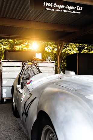 A vintage 1954 Cooper-Jaguar T33 car parked under a canopy with a warm sunset filtering through trees in the background. The car, with the number 34 displayed on its side, reflects the golden light, accentuating its sleek silver body. Tools and equipment are seen on a cabinet behind the car, suggesting a setting for maintenance or racing.