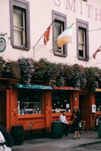 A street scene featuring a building with an orange facade and a sign reading 'T.P. Smiths'. There are flower baskets hanging above the windows, displaying a variety of colorful flowers. Two flags, one of which is the Irish flag, are mounted on poles attached to the building. Below the windows, a person is sitting on a barrel, and another person is walking past.
