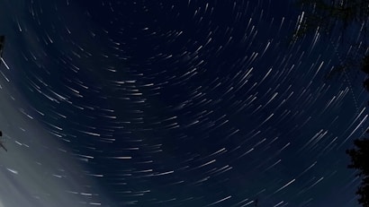 A night sky captured with long exposure photography, showing circular star trails against a deep blue background. There are silhouettes of trees and a structure visible at the edges, providing context to the natural scenery.