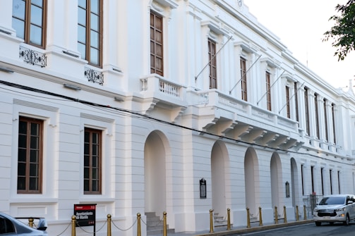 A large, elegant white building with classical architectural features including arches and decorative wrought iron accents above the windows. The building has multiple floors with tall, narrow windows and a series of flagpoles extending from the facade. A small sign in front reads 'Ayuntamiento de Manila'. Several cars are parked along the street, which is lined with gold-colored bollards connected by chains.