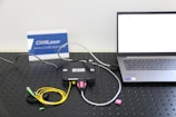 A setup involving a laptop connected to a black device with cables on a perforated black surface. There are colorful cables including yellow and green. A small pink object is placed nearby. A sign with the text 'CivilLaser' and a web address is in the background.