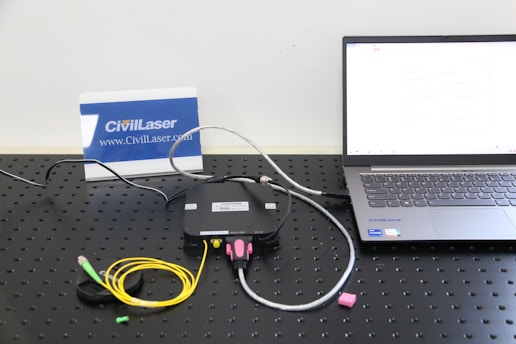 A setup involving a laptop connected to a black device with cables on a perforated black surface. There are colorful cables including yellow and green. A small pink object is placed nearby. A sign with the text 'CivilLaser' and a web address is in the background.