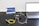 A setup involving a laptop connected to a black device with cables on a perforated black surface. There are colorful cables including yellow and green. A small pink object is placed nearby. A sign with the text 'CivilLaser' and a web address is in the background.