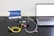 A setup involving a laptop connected to a black device with cables on a perforated black surface. There are colorful cables including yellow and green. A small pink object is placed nearby. A sign with the text 'CivilLaser' and a web address is in the background.