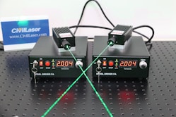 Two black DPPSS driver units with a digital display reading 2.004 are placed on a perforated surface. Each device is connected to a small module emitting a green laser beam. The setup includes various switches and indicators on the front panel, and there is a small CivilLaser card in the background displayed upright.