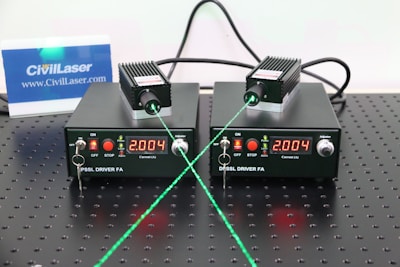 Two black DPPSS driver units with a digital display reading 2.004 are placed on a perforated surface. Each device is connected to a small module emitting a green laser beam. The setup includes various switches and indicators on the front panel, and there is a small CivilLaser card in the background displayed upright.