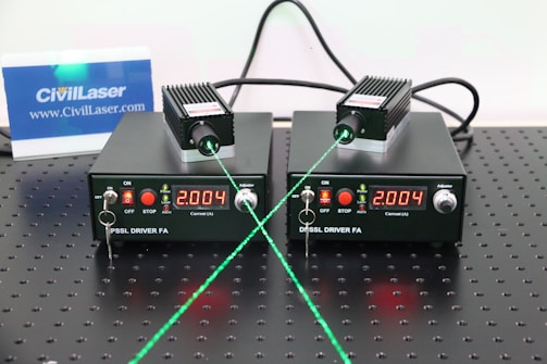 Two black DPPSS driver units with a digital display reading 2.004 are placed on a perforated surface. Each device is connected to a small module emitting a green laser beam. The setup includes various switches and indicators on the front panel, and there is a small CivilLaser card in the background displayed upright.