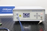 An optical fiber amplifier device sits on a perforated black metal surface. The device has a digital display showing numerical information and control buttons next to it. A red light indicator labeled 'ACTIVE' is illuminated. A key is inserted into a lock on the front panel. Blue fiber optic cables are connected to the IN and OUT ports.