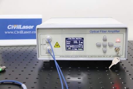 An optical fiber amplifier device sits on a perforated black metal surface. The device has a digital display showing numerical information and control buttons next to it. A red light indicator labeled 'ACTIVE' is illuminated. A key is inserted into a lock on the front panel. Blue fiber optic cables are connected to the IN and OUT ports.