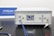 An optical fiber amplifier device sits on a perforated black metal surface. The device has a digital display showing numerical information and control buttons next to it. A red light indicator labeled 'ACTIVE' is illuminated. A key is inserted into a lock on the front panel. Blue fiber optic cables are connected to the IN and OUT ports.