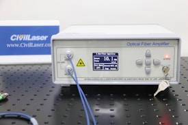 An optical fiber amplifier device sits on a perforated black metal surface. The device has a digital display showing numerical information and control buttons next to it. A red light indicator labeled 'ACTIVE' is illuminated. A key is inserted into a lock on the front panel. Blue fiber optic cables are connected to the IN and OUT ports.