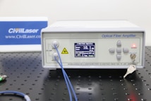 An optical fiber amplifier device sits on a perforated black metal surface. The device has a digital display showing numerical information and control buttons next to it. A red light indicator labeled 'ACTIVE' is illuminated. A key is inserted into a lock on the front panel. Blue fiber optic cables are connected to the IN and OUT ports.