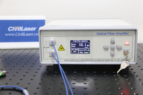 An optical fiber amplifier device sits on a perforated black metal surface. The device has a digital display showing numerical information and control buttons next to it. A red light indicator labeled 'ACTIVE' is illuminated. A key is inserted into a lock on the front panel. Blue fiber optic cables are connected to the IN and OUT ports.
