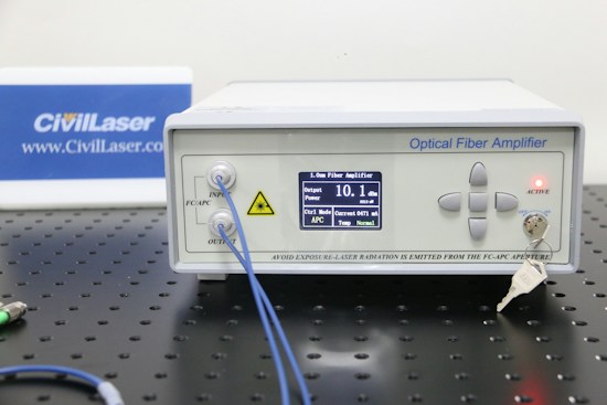 An optical fiber amplifier device sits on a perforated black metal surface. The device has a digital display showing numerical information and control buttons next to it. A red light indicator labeled 'ACTIVE' is illuminated. A key is inserted into a lock on the front panel. Blue fiber optic cables are connected to the IN and OUT ports.