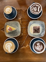 A wooden table is adorned with four cups of coffee artfully crafted with latte art, two of which are cappuccinos and two hot chocolates. There are also two dessert plates, one with a slice of cake topped with a strawberry and another with a piece of tiramisu. The table surface has a decorative carved pattern resembling a leaf.