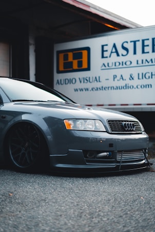 A sleek, modified Audi car with low suspension is parked in front of a building with a sign indicating 'Eastern Audio Limited'. The car is gray with a glossy finish, and the wheels are large and black, giving it a sporty appearance.
