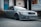 A sleek, gray Audi car is parked on a paved surface in an industrial setting. The car features aftermarket modifications, including a low stance and distinctive alloy wheels. In the background, there is a building with a blue door and a banner that reads 'Audio Visual'. The overall scene suggests a gritty, urban environment.