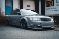 A sleek, gray Audi car is parked on a paved surface in an industrial setting. The car features aftermarket modifications, including a low stance and distinctive alloy wheels. In the background, there is a building with a blue door and a banner that reads 'Audio Visual'. The overall scene suggests a gritty, urban environment.