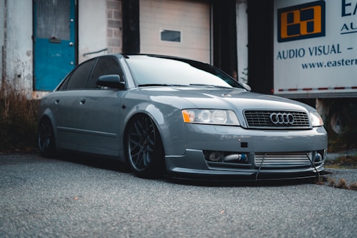 A sleek, gray Audi car is parked on a paved surface in an industrial setting. The car features aftermarket modifications, including a low stance and distinctive alloy wheels. In the background, there is a building with a blue door and a banner that reads 'Audio Visual'. The overall scene suggests a gritty, urban environment.