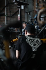 A person wearing a black shirt with an animal face design on the back is working on a bicycle. The scene appears to be in a workshop with various bike parts and tools visible. The environment is dimly lit, giving it a focused and industrious atmosphere.