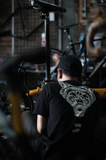 A person wearing a black shirt with an animal face design on the back is working on a bicycle. The scene appears to be in a workshop with various bike parts and tools visible. The environment is dimly lit, giving it a focused and industrious atmosphere.
