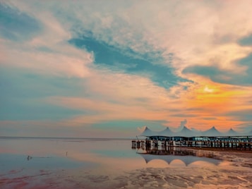 An expansive ocean view at sunset with a tranquil beach and a row of pavilions extending over the water. The sky is filled with vibrant colors ranging from oranges and yellows to soft blues and pinks, reflected in the calm sea surface. The beach in the foreground is slightly muddy, adding texture to the serene landscape.