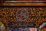 Intricate Arabic calligraphy is surrounded by ornate gold floral patterns on a dark background. The writing includes both circular and rectangular frames. The decorative style is rich and detailed, reflecting traditional artistry.