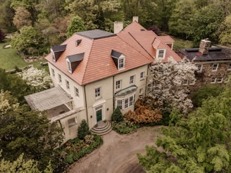An elegant two-story mansion with a distinctive red-tiled roof is surrounded by lush greenery and gardens. The house features multiple windows, a green entrance door, and a stone extension on the right. Trees and shrubs with various shades of green, white, and brown add to the serene ambiance.