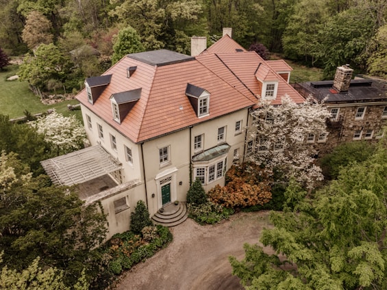 An elegant two-story mansion with a distinctive red-tiled roof is surrounded by lush greenery and gardens. The house features multiple windows, a green entrance door, and a stone extension on the right. Trees and shrubs with various shades of green, white, and brown add to the serene ambiance.