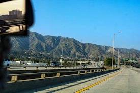 A highway scene with cars driving against a backdrop of mountain ranges. The road is wide and curves slightly to the right, bordered by concrete barriers. Traffic signs in green with white text are visible, and there are buildings and a reflection of a rearview mirror on the left side of the image.