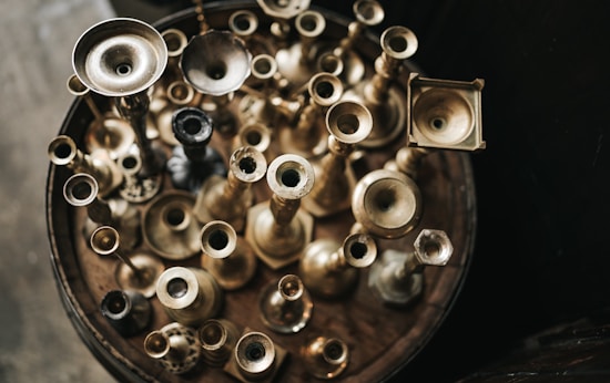 An assortment of vintage brass candlesticks is arranged closely together on a wooden surface, viewed from above. The varying sizes and shapes create an interesting visual pattern, with light reflecting off the different finishes of the candlesticks, adding depth and texture.
