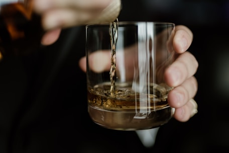 Close-up of a hand pouring clear spirit into a tasting glass on a wooden table.
