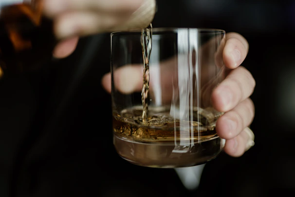 Elegant hands pouring aged whiskey from a decanter into a glass on a polished wooden table.