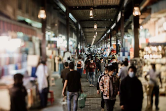 A bustling indoor market with numerous people walking along a corridor lined with shops. The area is well-lit with hanging lanterns, and various stalls displaying colorful fabrics and goods. Many individuals are wearing masks, indicating public health measures.