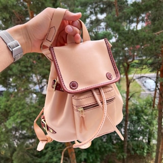 A hand is holding a small beige leather backpack with brown accents and stitching. The backpack features a front pocket with a zipper and a flap with decorative buttons. The background consists of lush green trees, indicating an outdoor setting.
