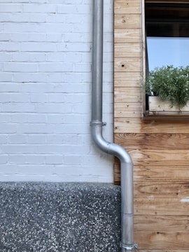 A brick and wood wall features a silver metal pipe and a window with a planter box filled with green plants. The bricks are painted white, contrasting with the natural wood tones. The base of the wall is made of textured concrete.