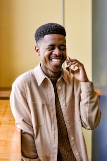 A person is smiling joyfully while talking on a mobile phone. They are leaning against a wooden surface and wearing a beige jacket over a textured brown shirt. The background consists of soft yellow and gray tones, creating a warm and relaxed ambiance.