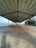 A large, open-air structure with a metal roof covering a concrete surface. Two yellow vehicles are parked under the roof, and a bright orange piece of equipment lies on the ground. Trees are visible in the background, and a shadow of a person is cast on the ground.