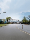 A wide road leads to a modern building with a glass facade. The sky above is clear and blue, while a few people walk near the entrance. Palm trees and manicured greenery line the path, creating a well-maintained and clean environment.