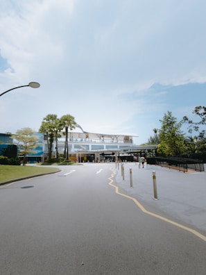 A wide road leads to a modern building with a glass facade. The sky above is clear and blue, while a few people walk near the entrance. Palm trees and manicured greenery line the path, creating a well-maintained and clean environment.