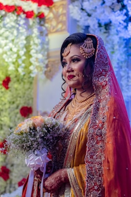 A bride is adorned in traditional wedding attire with intricate jewelry and a richly embroidered veil. She holds a bouquet of flowers featuring white and pale yellow roses. The background includes decorative floral arrangements in red and white.
