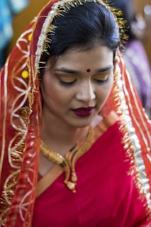 A woman is dressed in a vibrant traditional red saree with intricate golden embroidery. Her hair is neatly pinned up and partially covered with the saree's pallu. She wears bold makeup with red lipstick and golden eyeshadow, and a small red bindi adorns her forehead. A gold necklace is visible around her neck as she looks downward slightly.