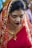 A woman is dressed in a vibrant traditional red saree with intricate golden embroidery. Her hair is neatly pinned up and partially covered with the saree's pallu. She wears bold makeup with red lipstick and golden eyeshadow, and a small red bindi adorns her forehead. A gold necklace is visible around her neck as she looks downward slightly.