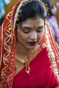 A woman is dressed in a vibrant traditional red saree with intricate golden embroidery. Her hair is neatly pinned up and partially covered with the saree's pallu. She wears bold makeup with red lipstick and golden eyeshadow, and a small red bindi adorns her forehead. A gold necklace is visible around her neck as she looks downward slightly.