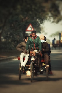 A man is pedaling a rickshaw on a road with three passengers seated behind him. The passengers are dressed in winter clothing, including a jacket, a hat, and a mask. The background features blurry trees and a traffic sign indicating a turn. The focus is on the group riding the rickshaw, with a soft, blurred landscape surrounding them.