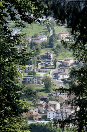 A view through the trees reveals a small village nestled in a lush green landscape. The village consists of a mix of residential houses with various architectural styles, surrounded by well-maintained gardens and grass. The winding road connects the homes, and cars are parked along the streets. The image captures a peaceful and serene environment, with dense foliage framing the aerial perspective of the village.