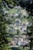 A view through the trees reveals a small village nestled in a lush green landscape. The village consists of a mix of residential houses with various architectural styles, surrounded by well-maintained gardens and grass. The winding road connects the homes, and cars are parked along the streets. The image captures a peaceful and serene environment, with dense foliage framing the aerial perspective of the village.