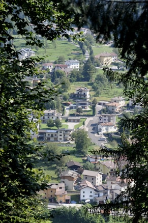 A view through the trees reveals a small village nestled in a lush green landscape. The village consists of a mix of residential houses with various architectural styles, surrounded by well-maintained gardens and grass. The winding road connects the homes, and cars are parked along the streets. The image captures a peaceful and serene environment, with dense foliage framing the aerial perspective of the village.