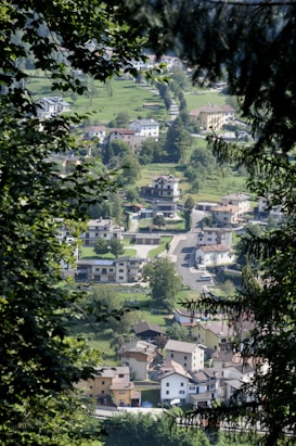 A view through the trees reveals a small village nestled in a lush green landscape. The village consists of a mix of residential houses with various architectural styles, surrounded by well-maintained gardens and grass. The winding road connects the homes, and cars are parked along the streets. The image captures a peaceful and serene environment, with dense foliage framing the aerial perspective of the village.