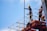 Two construction workers stand on a red metal scaffolding structure. They are wearing blue shirts and yellow safety helmets, suggesting an environment focused on safety and construction. One worker is holding onto a long metal rod as they both look into the distance under a clear blue sky.